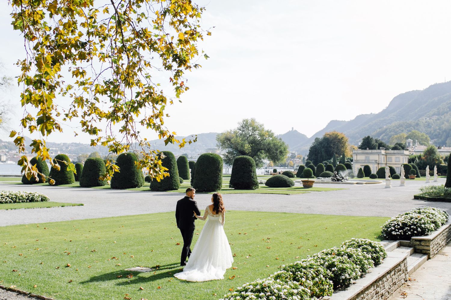 Para młoda spacerująca po eleganckim ogrodzie dworskim z widokiem na jezioro i rzeźby – romantyczna sceneria na wesele w dworku.